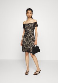 Vestido preto de renda com decote ombro a ombro, silhueta ajustada e bainha festonada. Modelo segura uma carteira estruturada preta e usa sandálias de tiras.