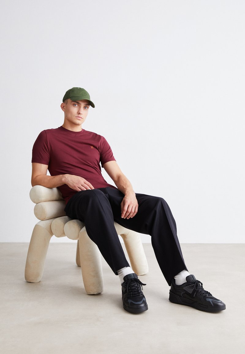 Bordowy t-shirt z krótkim rękawem z delikatnym logo, czarne spodnie i czarne sneakersy. Siedzi na kremowej, zaokrąglonej rzeźbie krzesła.