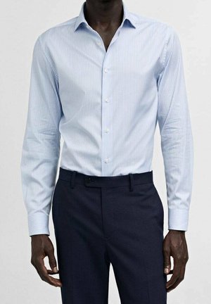 Mann trägt ein hellblau gestreiftes Langarmhemd, das in dunkelblaue, maßgeschneiderte Hosen gesteckt ist, und steht vor einem einfarbigen Hintergrund.