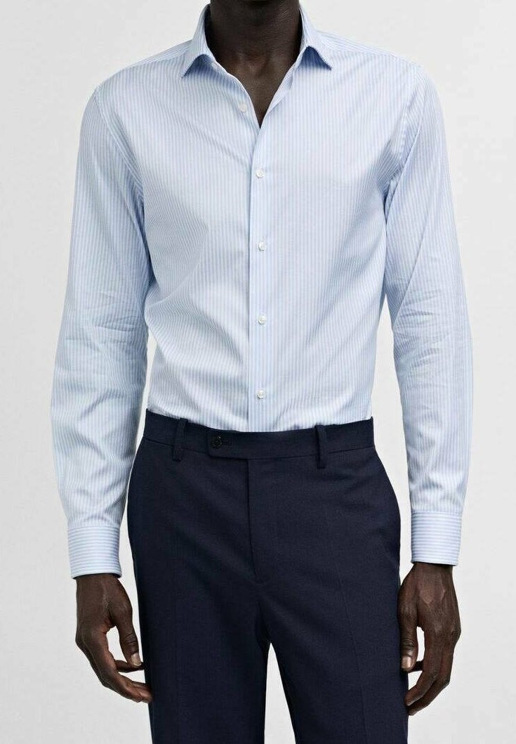 Homme portant une chemise à manches longues rayée bleu clair, rentrée dans un pantalon sur mesure bleu marine foncé, debout devant un fond uni.