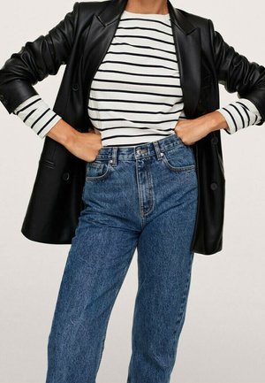 Femme portant un jean bleu taille haute, un t-shirt à manches longues blanc et noir à rayures, et un blazer en cuir noir, mains sur les hanches.