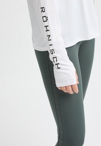 Weißes Langarmshirt mit schwarzem "Röhnisch"-Logo am Ärmel, kombiniert mit dunkelgrünen High-Waist-Leggings, die einen dezenten Logo-Print an der Seite aufweisen.