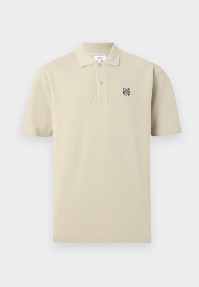 Μπεζ κοντομάνικο πόλο με δύο κουμπιά και μικρό κεντημένο λογότυπο με κεφάλι λύκου στην αριστερή πλευρά του στήθους.