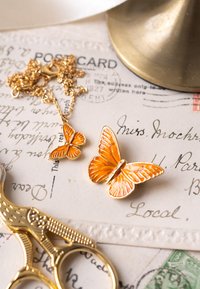 Auksinės spalvos drugelių papuošalų rinkinys, apimantis didelį ir mažesnį drugelį, abudu su oranžinės emalės detalėmis, padėti ant senovinių atvirukų ir žirklių.