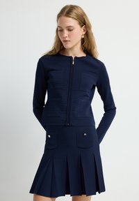 Chaqueta texturizada de color azul marino con mangas largas y cremallera frontal, combinada con una falda plisada que cuenta con bolsillos laterales y acentos de botones dorados.