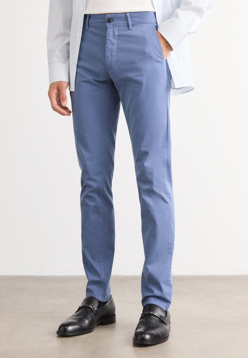 Pantalons en coton bleu clair avec une coupe slim, un devant plat et des poches latérales, associés à des chaussures en cuir noir avec un accent de boucle.