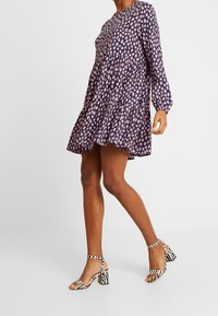 Lila blommig klänning med långa ärmar och samlad midja, matchad med svarta och vita randiga klacksandaler. Texturerad tygdetalj synlig.