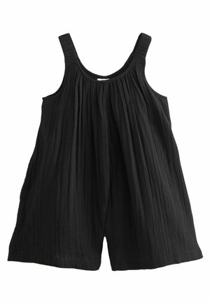 Enfant portant une robe noire sans manches et des sandales dorées, debout avec une jambe étendue, fond beige avec un grand coquillage à proximité.