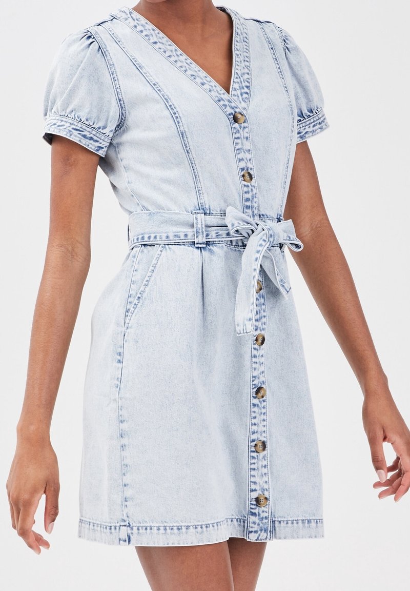 Light blue denim dress with short puff sleeves, a V-neck, front buttons, and a fabric belt at the waist. Distressed texture and stitching details.