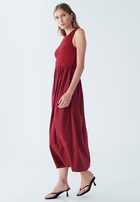 Rdeča maksi obleka z nabranim korzetom, tekočo krilno in enim naramnim trakom. Nošena s črnimi sandali z več trakovi. Gladek tekstura.