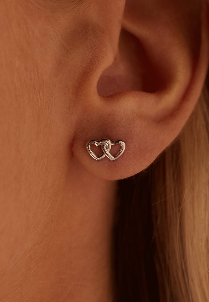 Clou d’oreille en argent en forme de deux cœurs entrelacés porté sur un lobe d'oreille à peau claire.