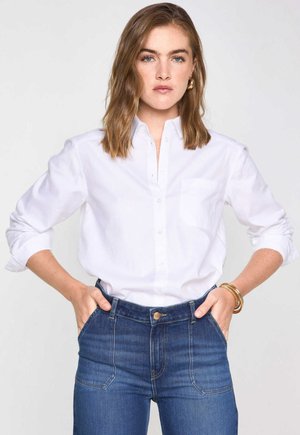 Donna con capelli castani fino alle spalle che indossa una camicia bianca con bottoni e maniche arrotolate, jeans blu a vita alta e gioielli d'oro.