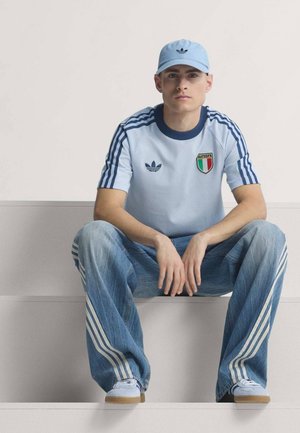 Ung mand siddende iført lyseblå Adidas kasket, lyseblå italiensk sportstrøje, løse jeans med hvide striber og hvide sneakers.