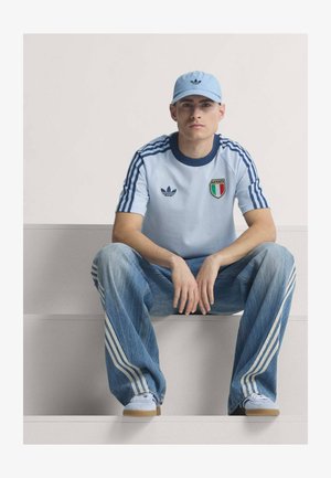 Ung mand siddende iført lyseblå Adidas kasket, lyseblå italiensk sportstrøje, løse jeans med hvide striber og hvide sneakers.