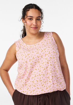 Kobieta o ciemnych, kręconych włosach związanych do tyłu, ubrana w różowy top bez rękawów z drobnym żółtym kwiatowym wzorem oraz ciemnobrązowe spodnie, stojąca z rękami w kieszeniach.