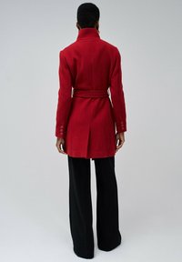 Abrigo rojo de mezcla de lana con cuello alto, cintura con cinturón y puños abotonados, combinado con pantalones negros de pierna ancha. Textura suave, ajuste entallado.