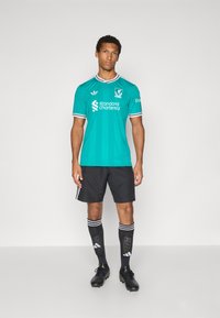 Teal målmannstrøye med svarte og hvite stripete detaljer, kombinert med svarte shorts og stripete knestrømper, båret med svarte fotballsko.