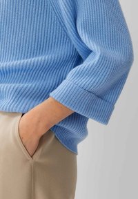 Pull en maille côtelée bleu clair avec des poignets repliés, associé à un pantalon beige. La texture est douce et les coutures sont soigneusement finies.