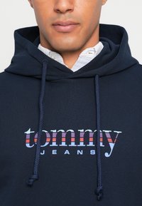 Navy hoodie with drawstrings, featuring a printed logo reading "tommy JEANS" in white and striped accents in red and blue. Smooth texture.