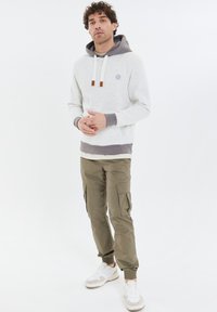 Felpa con cappuccio grigia chiara, con cappuccio interno grigio, coulisse e dettagli marroni. Pantaloni cargo verde oliva con tasche laterali e polsini elasticizzati. Sneakers bianche.