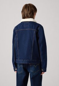 Giacca di jeans di colore blu scuro con colletto foderato in pile, caratterizzata da cuciture a contrasto e polsini con bottoni. Il retro presenta una pattina centrale e un'etichetta.