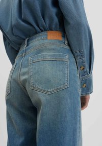 Des jeans en denim bleu clair présentent une taille haute, cinq poches et une application rectangulaire en cuir beige sur la ceinture arrière. Détails en boutons sur les manches.