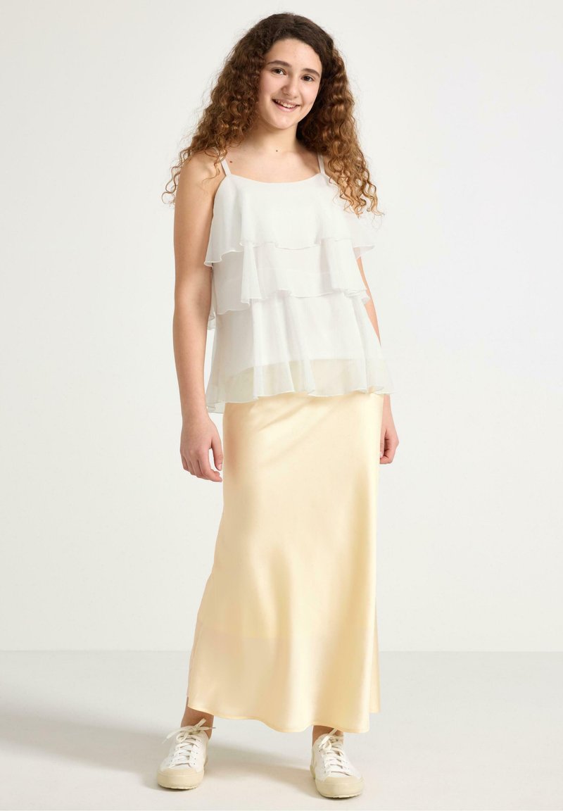 Top in chiffon bianco a strati con spalline regolabili, abbinato a una gonna maxi in satin giallo pallido e sneakers bianche. Texture liscia e design fluido.