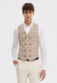 Beżowa kamizelka w kratę z podwójnym zapięciem, posiadająca pięć guzików, dwie przednie kieszenie oraz fakturowany materiał, zestawiona z białą koszulą.
