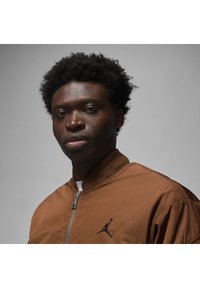 Giacca bomber marrone con chiusura a zip, colletto rigato e logo nero di Jordan sul petto. Tessuto liscio e leggero, su uno sfondo neutro.