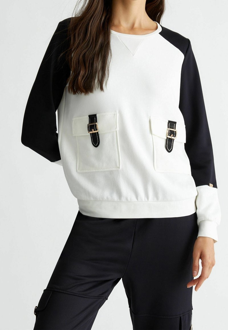 Femme portant un haut à manches longues blanc et noir avec deux poches avant munies de sangles à boucle noire et un pantalon noir.