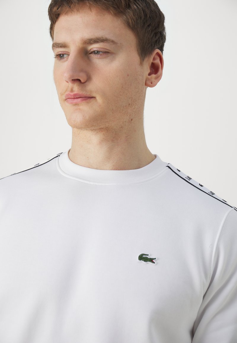 Felpa bianca con colletto a girocollo, con accenti neri sui lati e un piccolo logo ricamato a forma di coccodrillo verde sul petto. Tessuto morbido.