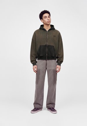 Jeune homme debout portant un sweat à capuche zippé vert foncé, un pantalon gris à jambes larges et des baskets violettes sur un fond clair uni.