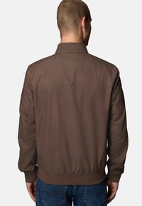 Veste aviateur marron avec une texture lisse, des poignets et un ourlet côtelés. Présente un design simple et classique ; portée sur un jean en denim.