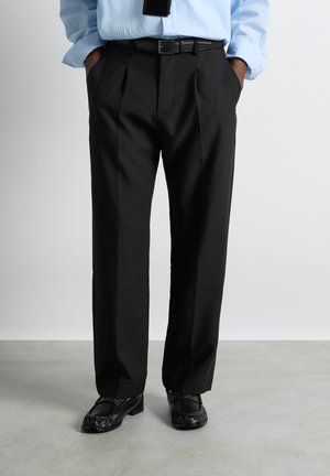 Muž nosící černé plisované kalhoty, černý pásek, černé mokasíny a modrou pruhovanou košili s rukama v kapsách.