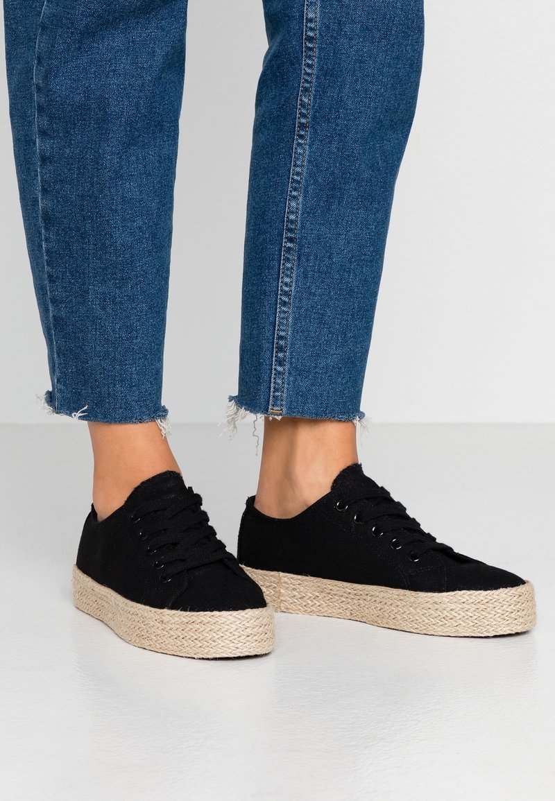 Jambes portant un jean bleu à ourlet effiloché et des baskets à plateforme noires à lacets avec semelles tissées beiges sur un sol clair.