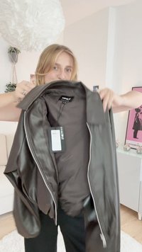 Black faux leather jacket with shiny finish, wide collar, and zip closure, showing inner lining and care tag on a model's hand.