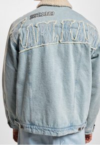 Chaqueta de denim azul clara con cuello de borrego, texto desgastado "KARL KANI" y "EST. 1989" bordado en la parte posterior.