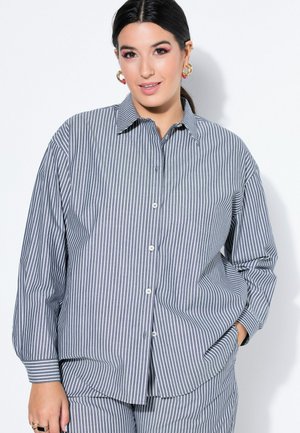 Frau mit dunklen Haaren, die goldene Creolen und ein blau-weiß vertikal gestreiftes Hemd mit Knöpfen sowie passende Hosen trägt.