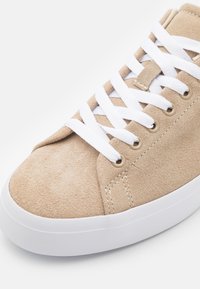 Sneaker in suede beige con lacci bianchi, caratterizzata da occhielli rotondi e una suola in gomma bianca. Texture liscia e dettagli di design minimi.