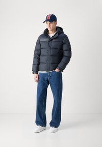 Giacca a vento scura con cappuccio e chiusura a bottoni, abbinata a jeans blu larghi e sneaker bianche. Il modello indossa un cappellino blu navy con strisce rosse.