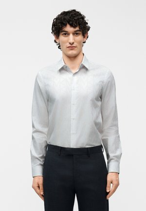 Muž s kučeravými vlasmi, oblečený v bielej vzorovanej košeli s dlhým rukávom a tmavých nohaviciach, stojaci pred jednoduchým pozadím.