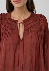 Lang, rode blouse met een gerimpelde halslijn, geplooide textuur en striksluiting. Heeft een sleutelgatopening en vloeiende mouwen.