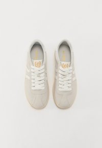 Beige Wildledersneakers mit weißen Schnürsenkeln, runder Spitze, strukturierten Sohlen und dezenten goldenen Logodetails auf der Zunge und den Seitenstreifen.