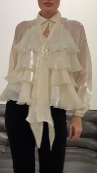 Γυναίκα που φοράει μια διάφανη, μπεζ μπλούζα με στρωματοποιημένες βολάν και δεμένη φουλάρι στον λαιμό, συνδυασμένη με μαύρο παντελόνι, στέκεται σε εσωτερικό χώρο.