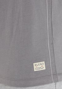 Szürke pamut póló, testhez simuló szabással, fehér varrással és „BLEND 1993 DENIM WITH ATTITUDE” feliratos címkével az alsó szegélyén.
