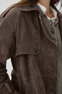 Lähikuva henkilöstä, joka käyttää ruskeaa mokkanahkatrenssiä harmaan neulotun neuletakin ja valkoisen kauluspaitaan päälle.