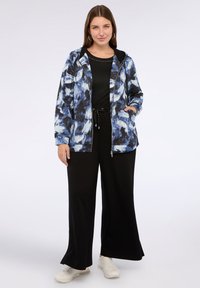 Giacca zip con cappuccio a motivo astratto blu e bianco, abbinata a una maglietta nera e pantaloni larghi neri. Sneakers bianche completano il look.
