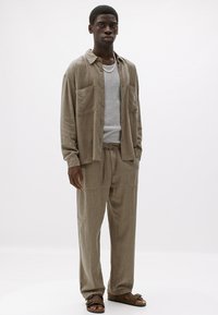 Cămașă bej din catifea cu două buzunare, tricou gri cu guler rotund, pantaloni bej asortați și sandale maro. Textură netedă, croi lejer.
