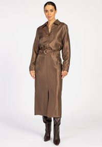 Vrouw in een glanzende bruine midi-jurk met riem, een rits aan de voorkant en lange mouwen, gecombineerd met donkere knie-laarzen, staande tegen een witte achtergrond.