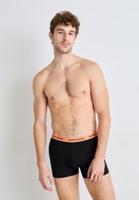 Black men's boxer shorts with an orange elastic waistband featuring "BJÖRN BORG" logo. Smooth fabric, fitted design, knee-length style.